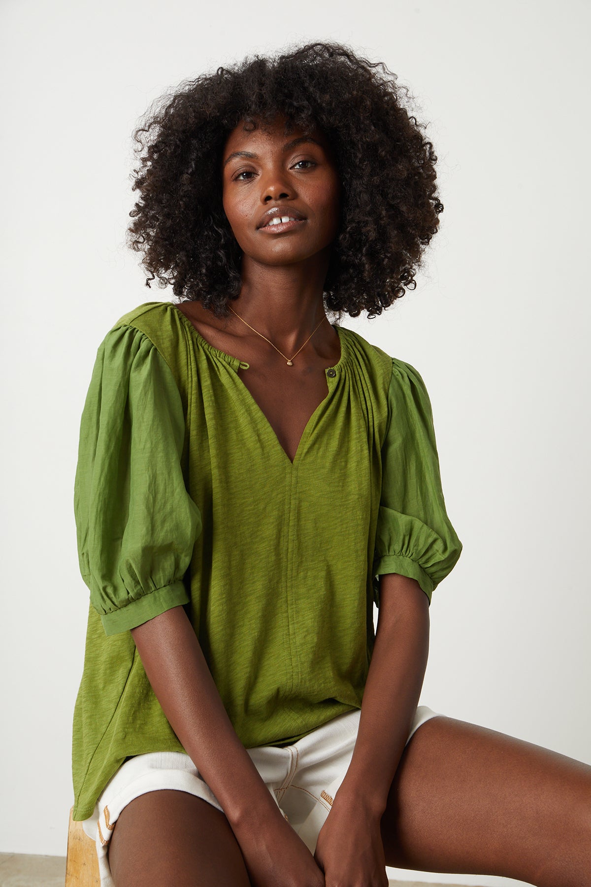   Woman sitting on stool in studio wearing Mallory Top in moss green with puff sleeves paired with white denim shorts front 
