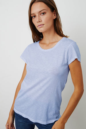 A person with long hair is wearing a light blue, short-sleeve ODELIA COTTON SLUB CREW NECK TEE by Velvet by Graham & Spencer and jeans, standing against a plain white background.