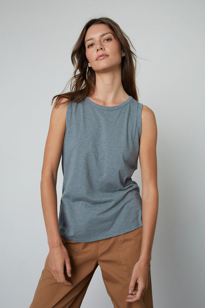 A woman with long brown hair wearing a sleeveless light blue TAURUS TANK TOP by Velvet by Graham & Spencer and light brown pants, a versatile staple in any wardrobe, stands against a plain background.