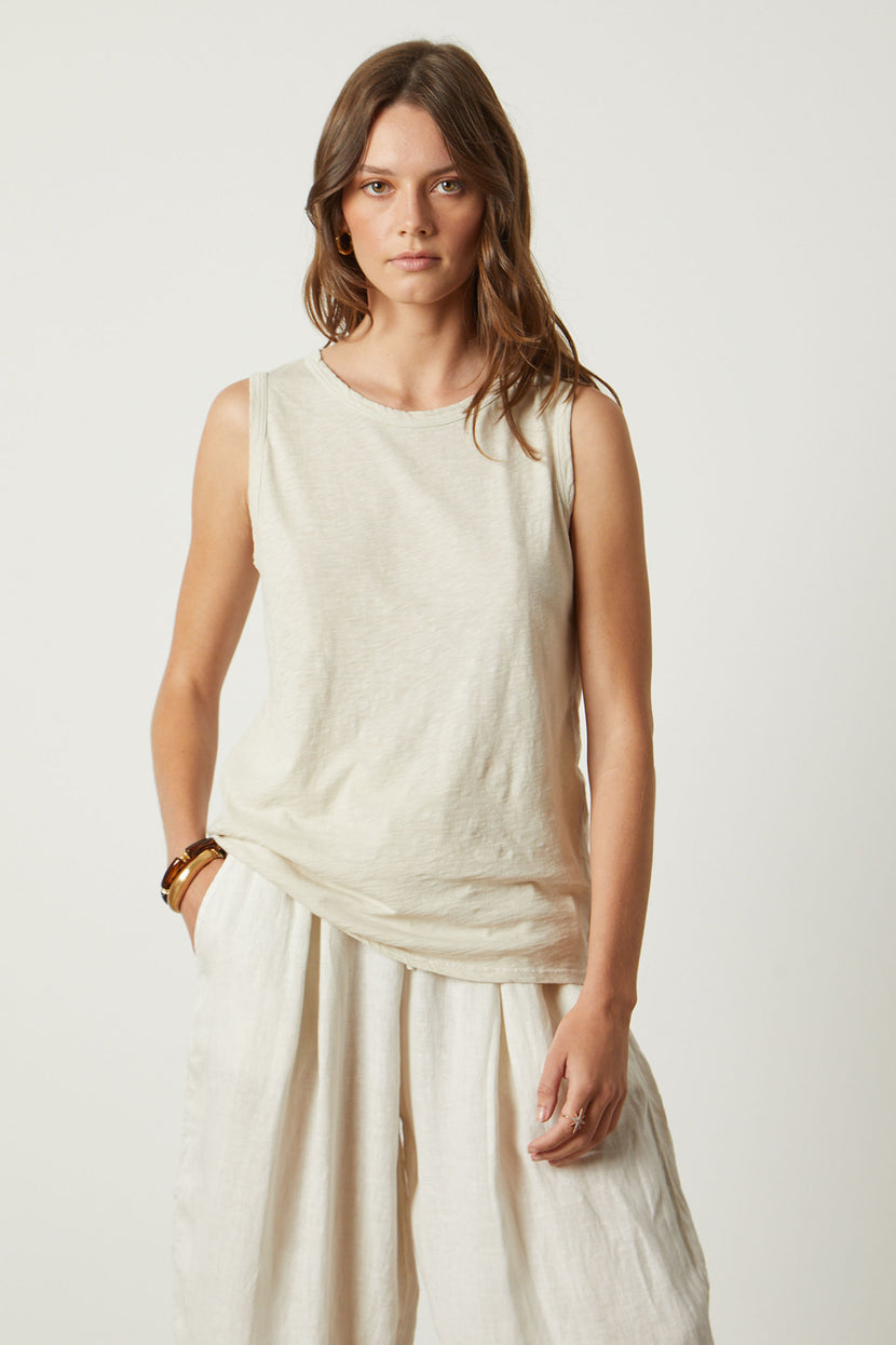 A woman wearing a sleeveless beige TAURUS TANK TOP by Velvet by Graham & Spencer and loose white pants, crafted from textured cotton slub, stands against a plain background, looking directly at the camera.