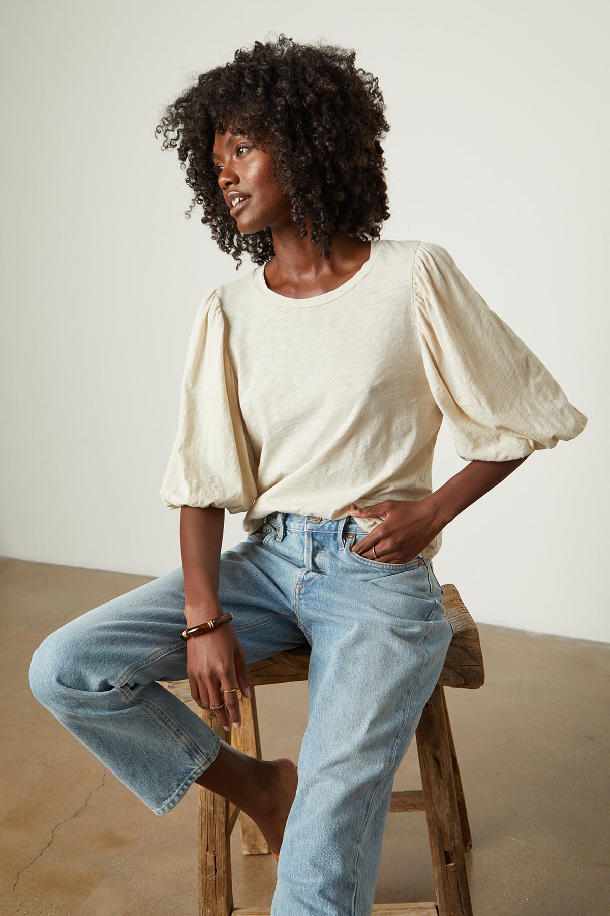   A woman is sitting on a stool wearing UMA PUFF SLEEVE TEE by Velvet by Graham & Spencer, jeans, and a blouse. 