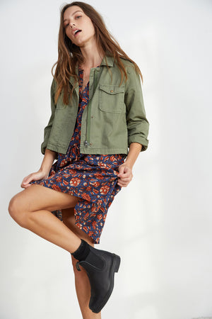 A woman wearing an olive green jacket over the SHOSHANA PRINTED DRESS by Velvet by Graham & Spencer strikes a pose, lifting one leg while looking upward. She is also wearing black ankle boots.