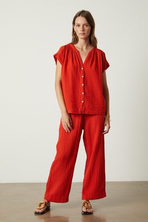 A woman in red pants and sandals is standing in front of a white wall wearing the Pamela Cotton Gauze Button-Up Top by Velvet by Graham & Spencer.
