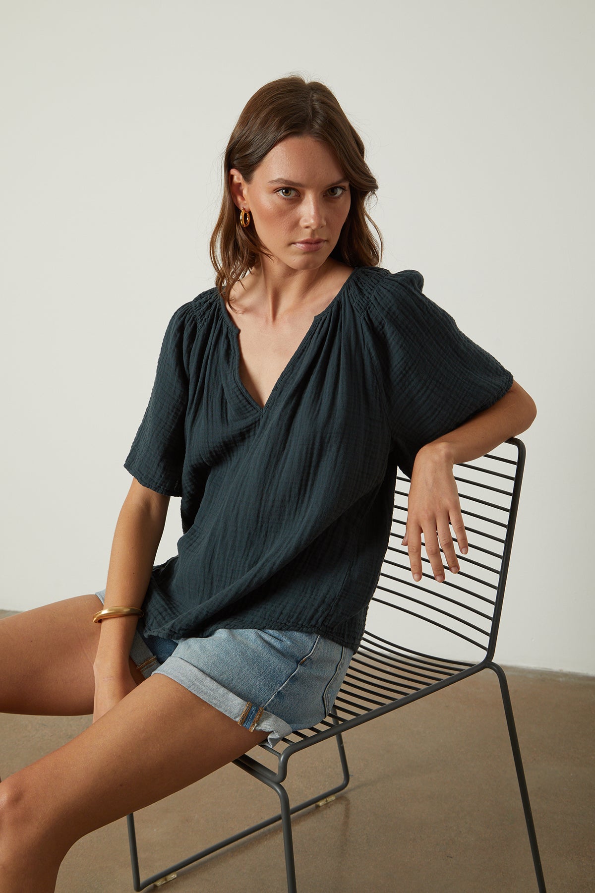 the model is sitting on a chair in a dark blue Velvet by Graham & Spencer SADIE COTTON GAUZE V-NECK TOP.-26022630981825