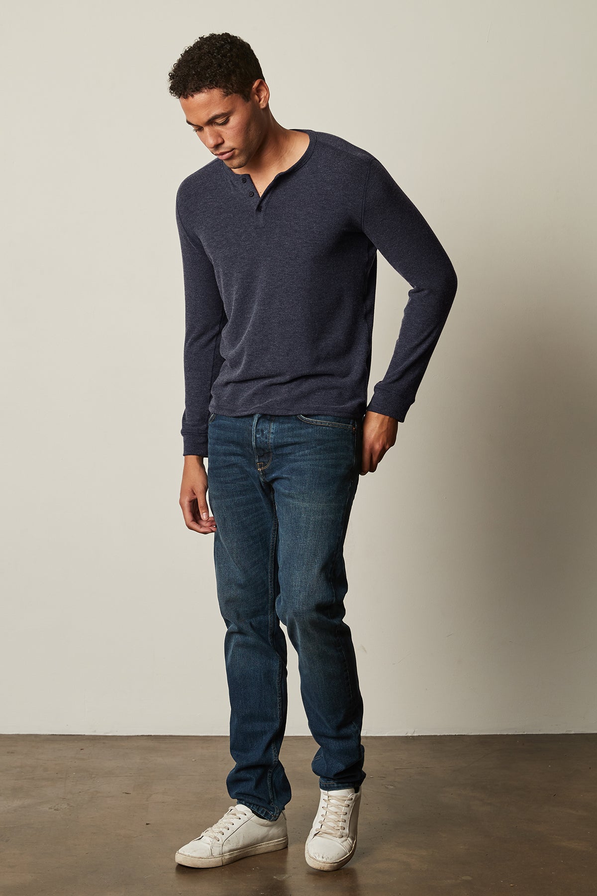 A versatile man, donning Velvet by Graham & Spencer jeans and a FAUST COZY JERSEY HENLEY sweater, confidently poses in front of a white wall.-25484011798721