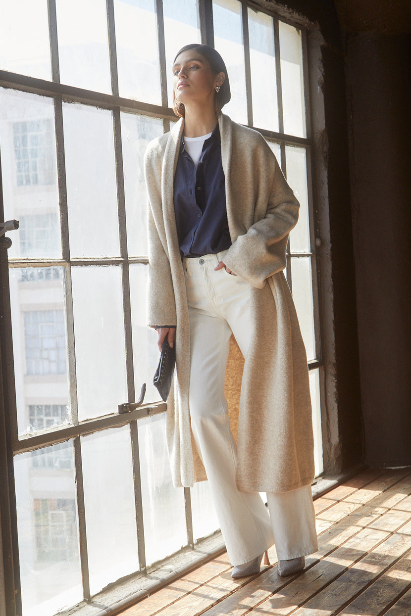 A woman in a Velvet by Jenny Graham Carmel Coat and white pants standing in front of a window.