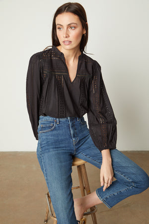 Model sitting on stool in studio wearing Jordyn Embroidered V-Neck Top in black tucked into blue denim front