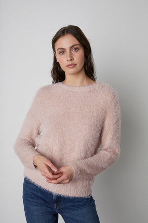 A woman wearing a light pink, kitten-soft stretchy knit RAY CREW NECK SWEATER by Velvet by Graham & Spencer and blue jeans stands against a plain background.