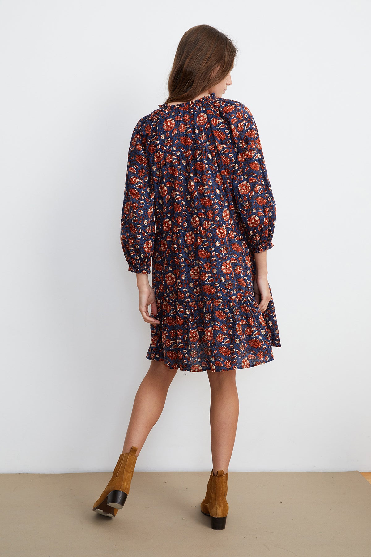 A person stands facing away, wearing the SHOSHANA PRINTED DRESS by Velvet by Graham & Spencer. The dark floral dress with long sleeves and the elegant ruffle skirt are complemented by brown ankle boots, all against a plain white background.-7835042185297