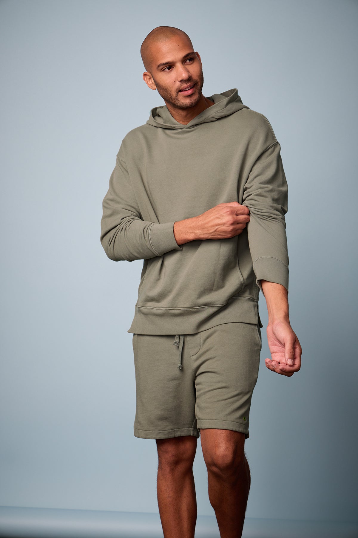   Lookbook image of Dan Hoodie in camp muted green french terry with Kane short, sleeves pushed up, blue background front 