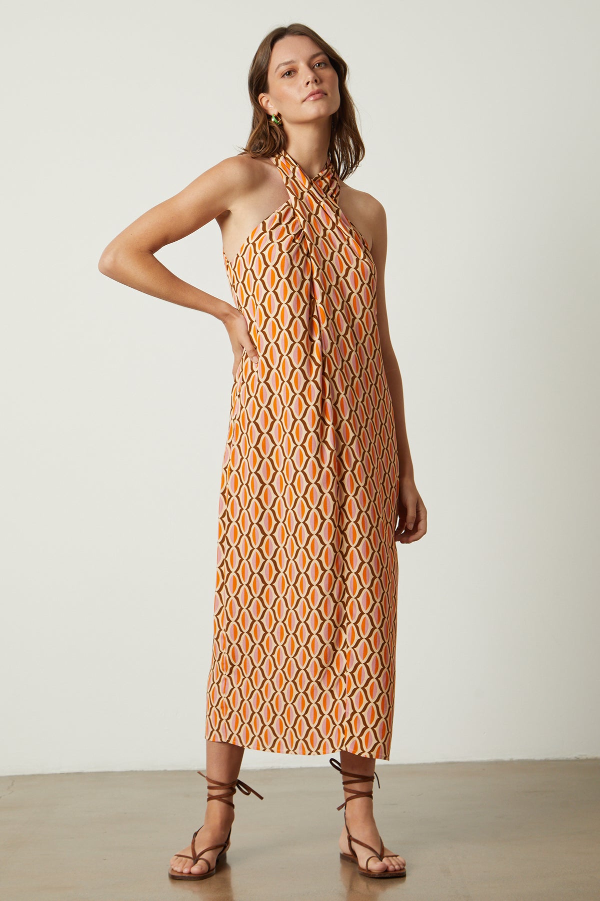 Women standing with hand on hip wearing Caterina dress in orange geometric pattern gold earrings and sandals full length front-26079026938049