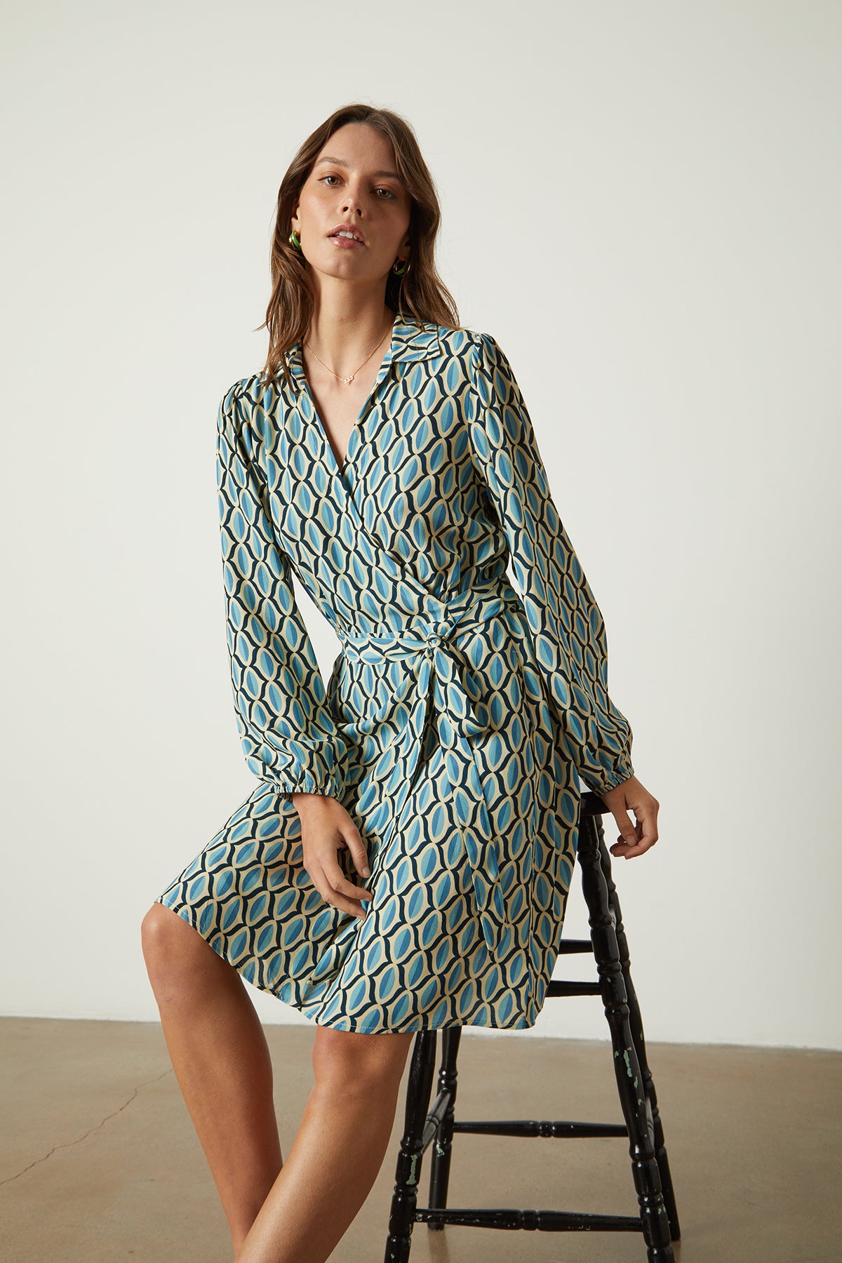 Model sitting on black wooden stool in studio wearing Gabriella wrap dress in blue geometric print crepe front-26079061082305