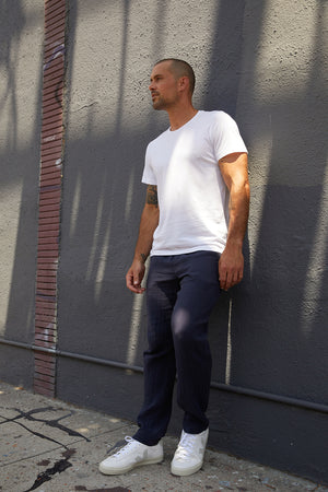 White Angelo crew neck graphic tee and navy ryan pants, model standing outside against wall.
