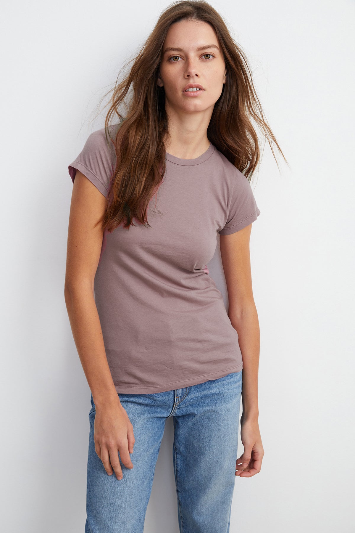 A woman with long brown hair, wearing a Velvet by Graham & Spencer JEMMA TEE and blue jeans, is standing against a plain white background, looking at the camera.-23854328283329