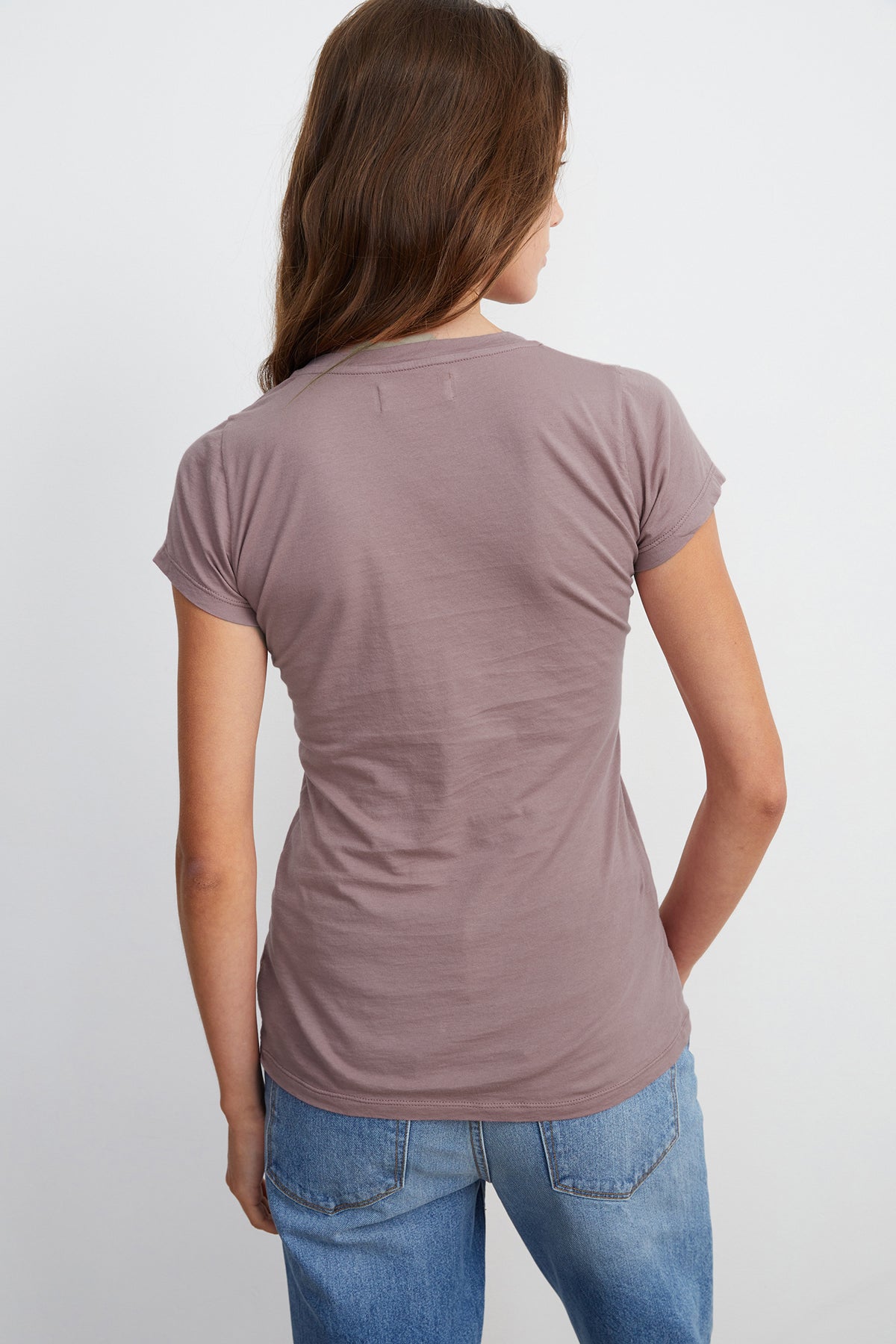   A person with long brown hair is seen from the back wearing a timeless staple: an ultra-soft gauzy whisper, mauve-colored Velvet by Graham & Spencer JEMMA TEE and blue jeans. 