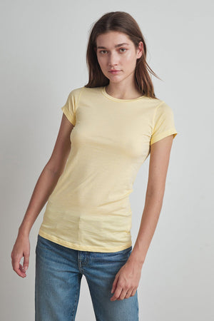 A woman with brown hair, wearing an ultra-soft JEMMA TEE by Velvet by Graham & Spencer and blue jeans, stands against a plain background, facing the camera.