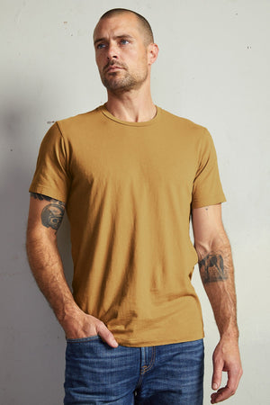 A man with a beard and tattoos on both arms stands against a plain background, wearing the HOWARD TEE by Velvet by Graham & Spencer, made from lightweight cotton knit that offers a perfect fit, paired with blue jeans.