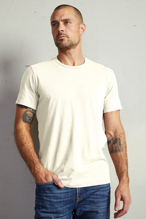 A man with short hair and tattoos on both arms is wearing a plain white, lightweight Velvet by Graham & Spencer HOWARD TEE and jeans. He has one hand in his pocket and is standing against a light-colored wall, showcasing the perfect fit of his attire.