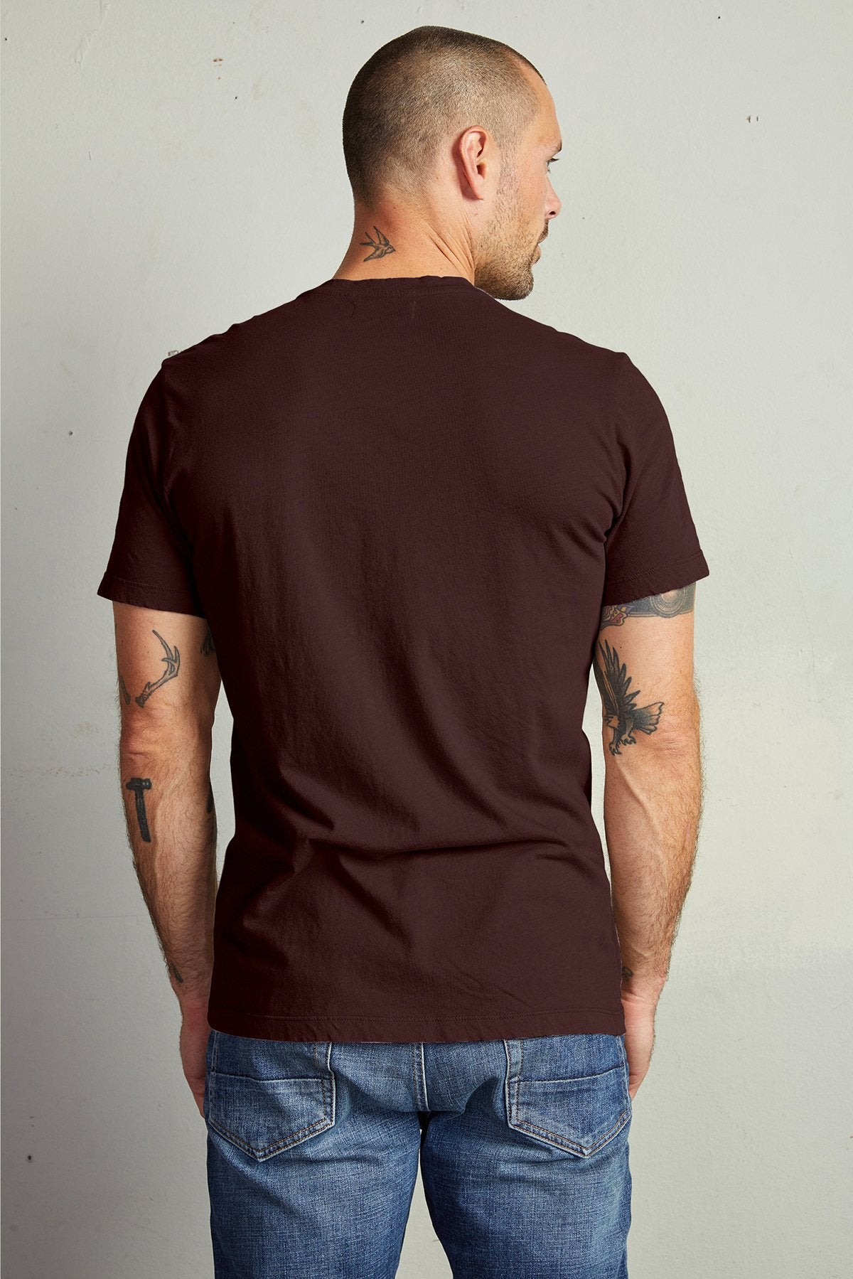   A man with a shaved head wearing a maroon Velvet by Graham & Spencer HOWARD TEE made of lightweight cotton knit and blue jeans stands facing away, displaying tattoos on his neck and arms. 