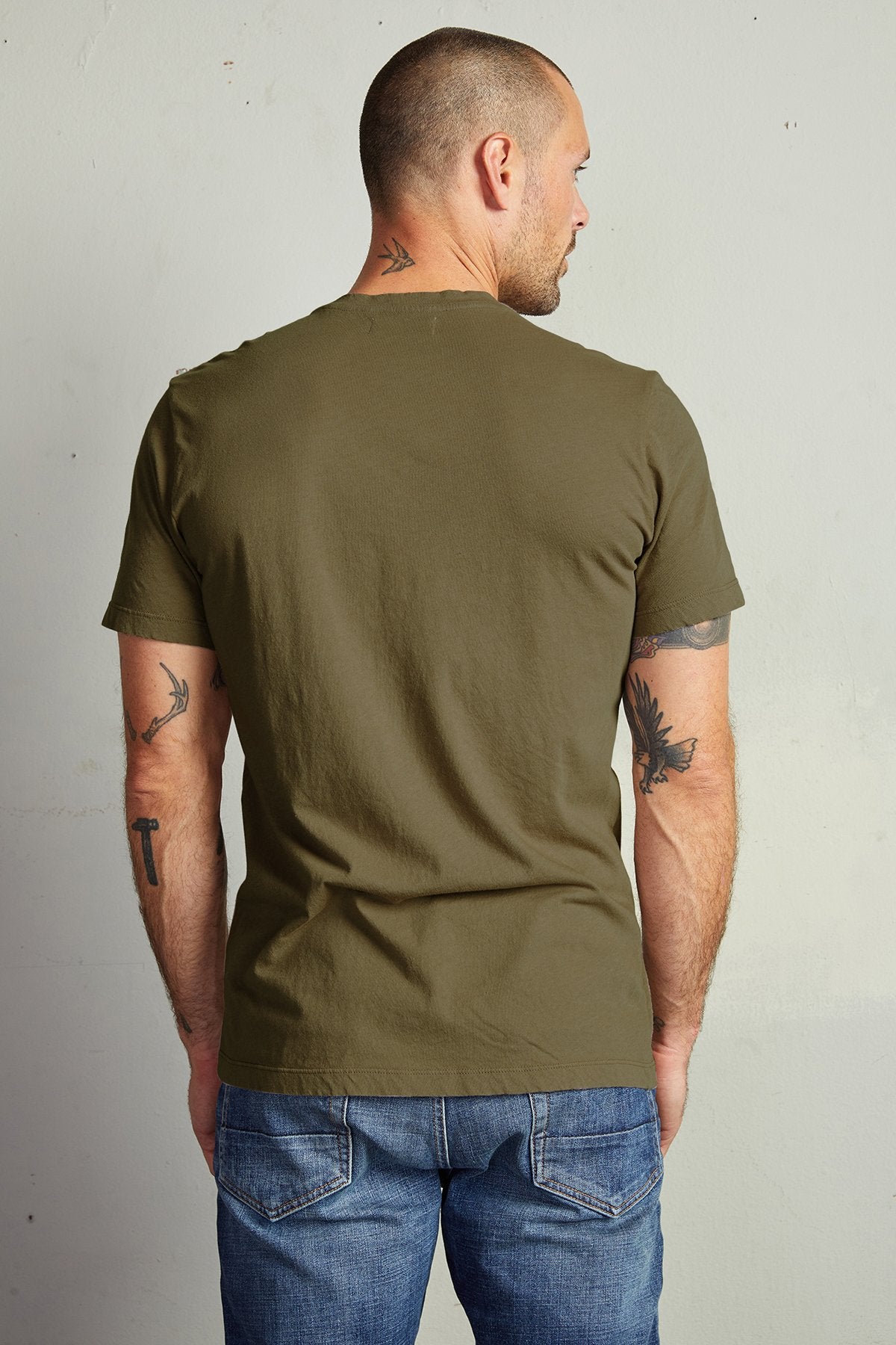 A bald man with tattoos stands with his back to the camera, wearing an olive green t-shirt made of lightweight cotton knit and blue jeans. He's donning the HOWARD TEE by Velvet by Graham & Spencer.-23687645692097