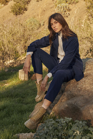 Model sitting outside on boulder wearing Jeslyn Corduroy Button-Up Shirt in dark blue night color, Renny Sweater and Aspen pants in navy. 