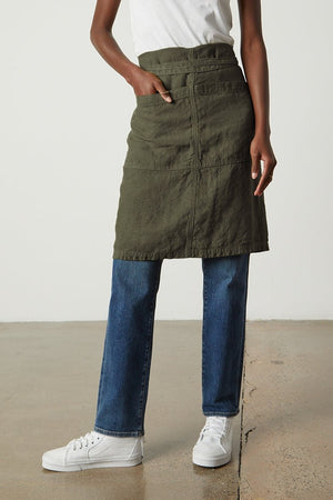 A person stands wearing a white t-shirt, an olive-green skirt over blue jeans, and white sneakers, with one hand in a pocket. They sport the Jenny Graham Home LINEN APRON with an adjustable neck strap, all against a plain background.