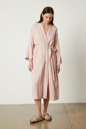 A woman wearing a pink cotton gauze robe and sandals from Jenny Graham Home.