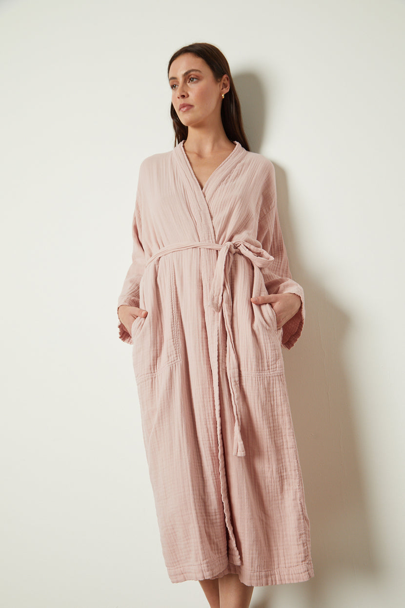 A woman wearing a Jenny Graham Home pink cotton gauze robe is standing against a wall.