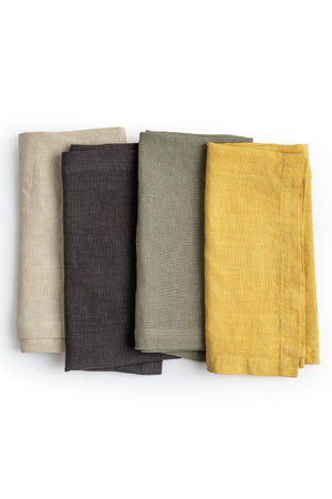 Four elegant Jenny Graham Home linen napkins in beige, charcoal, olive green, and mustard yellow, neatly folded and stacked on a white background.