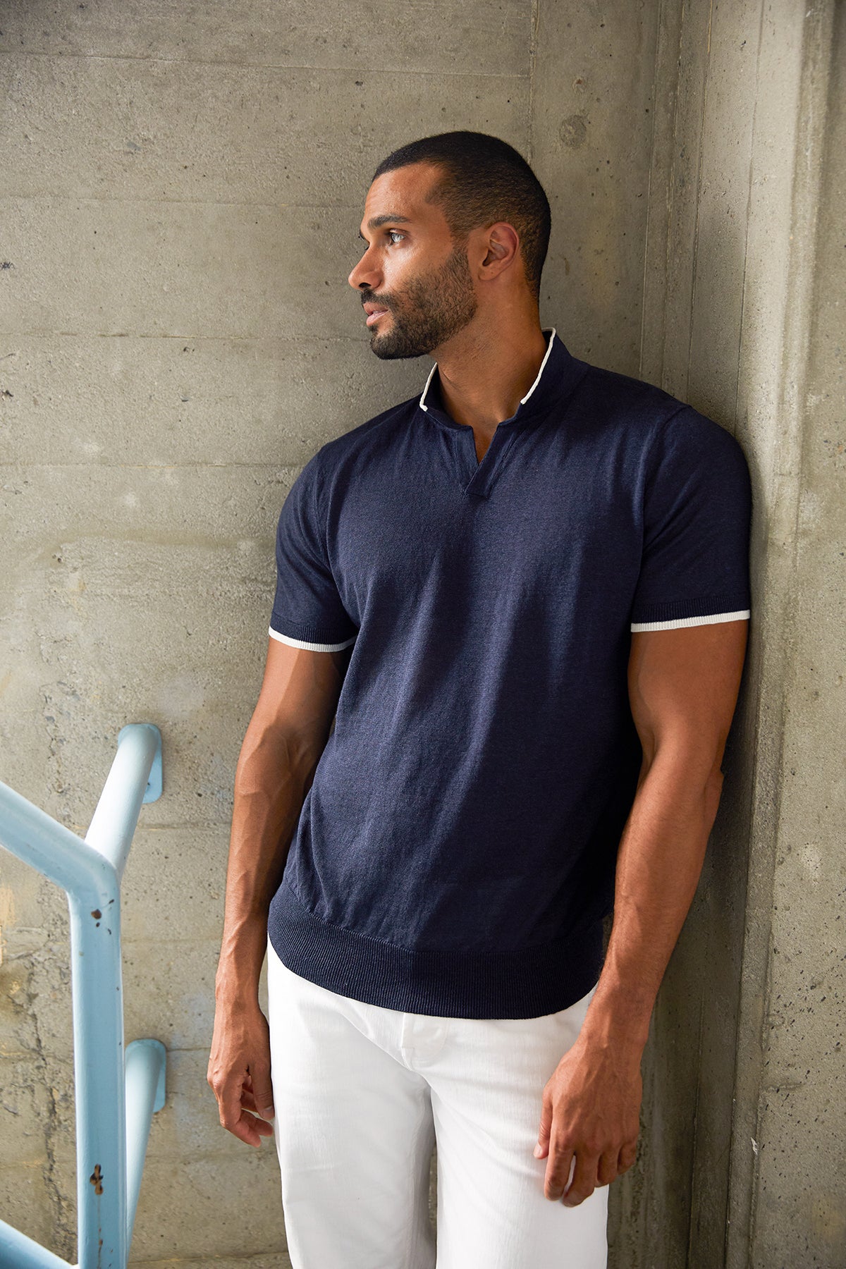   Navy blue Gordon linen blend polo shirt with white stripe trim around sleeves and at collar, with model leaning against wall looking to the side, wearing white denim. 