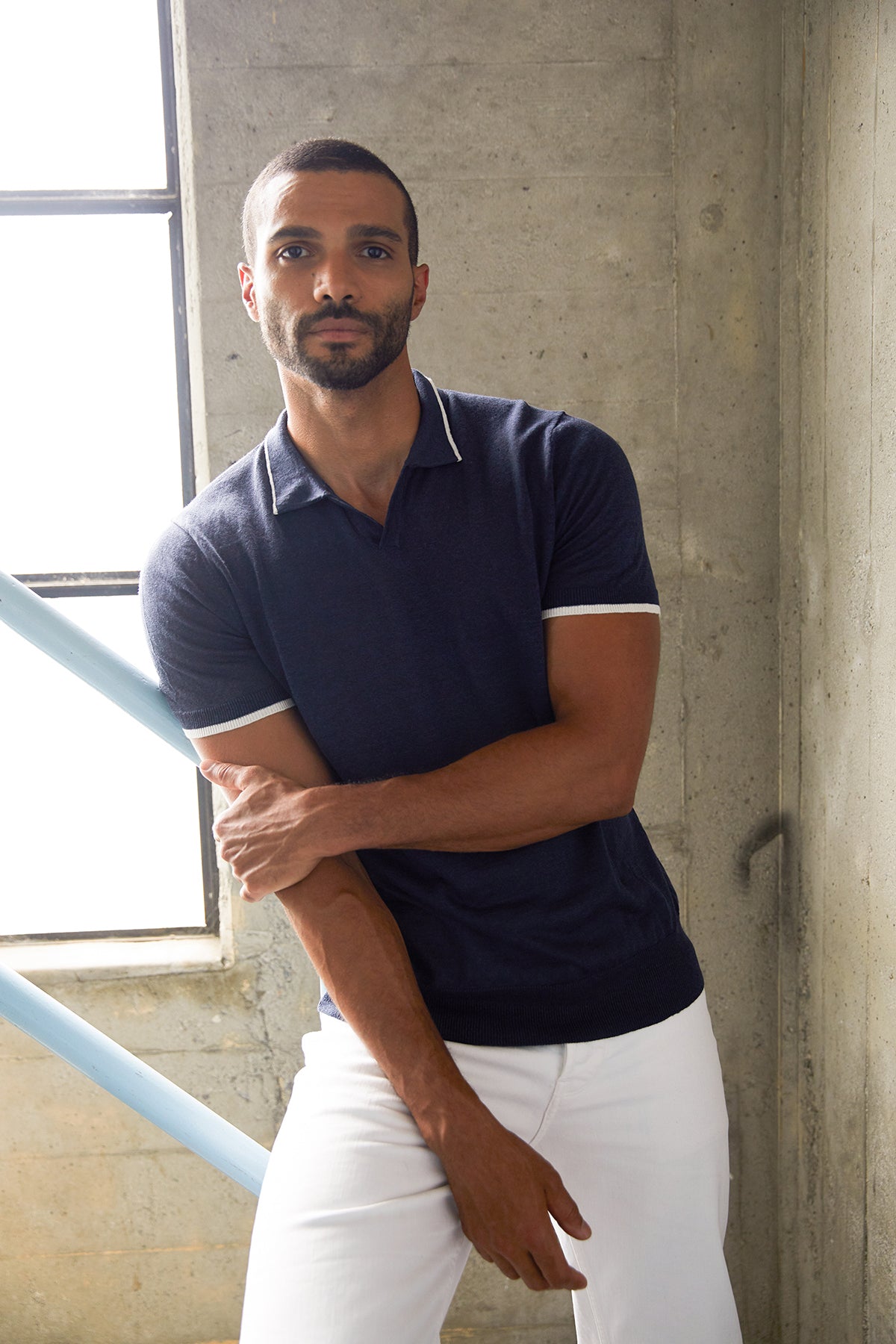   Model leaning on stair railing near window wearing navy blue Gordon linen blend polo shirt with white stripe trim around sleeves and at collar, with white denim. 