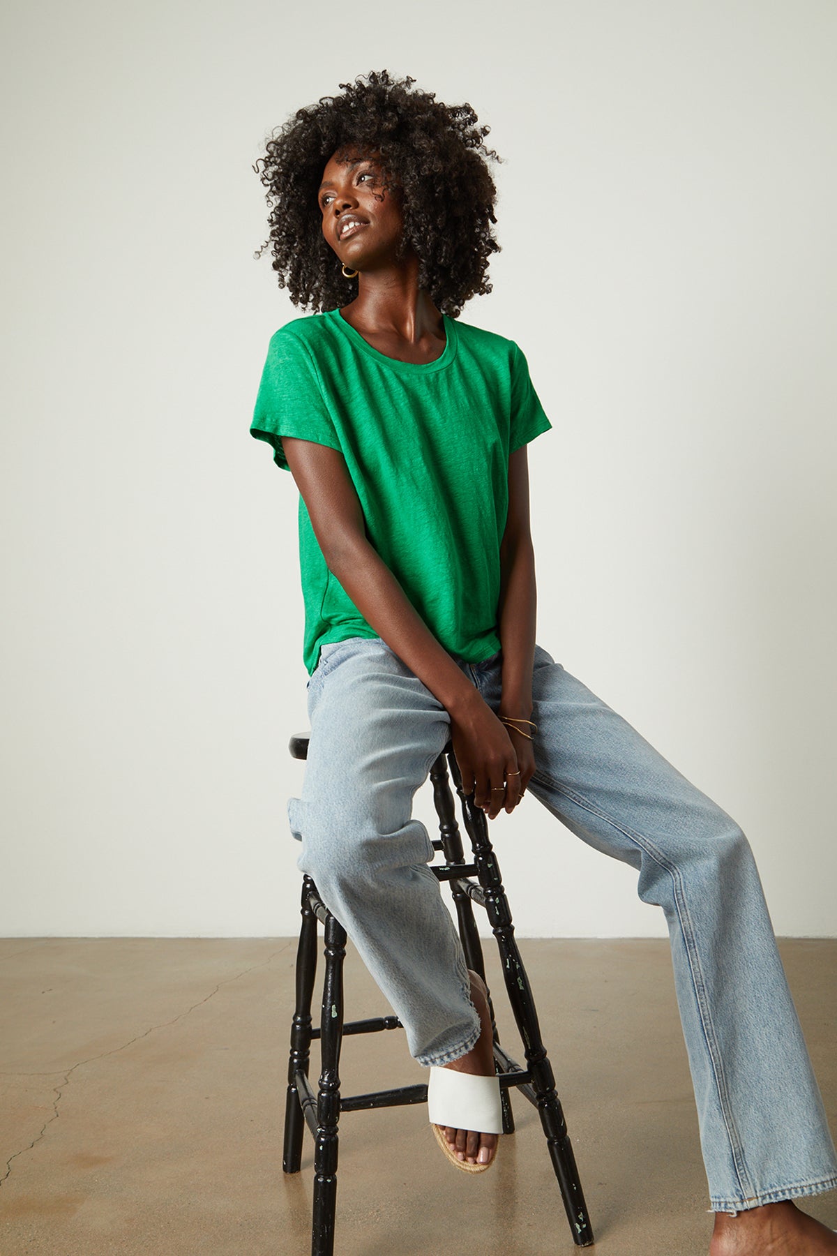   Model sitting on stool in studio wearing Casey Tee in emerald with blue denim and white slides 