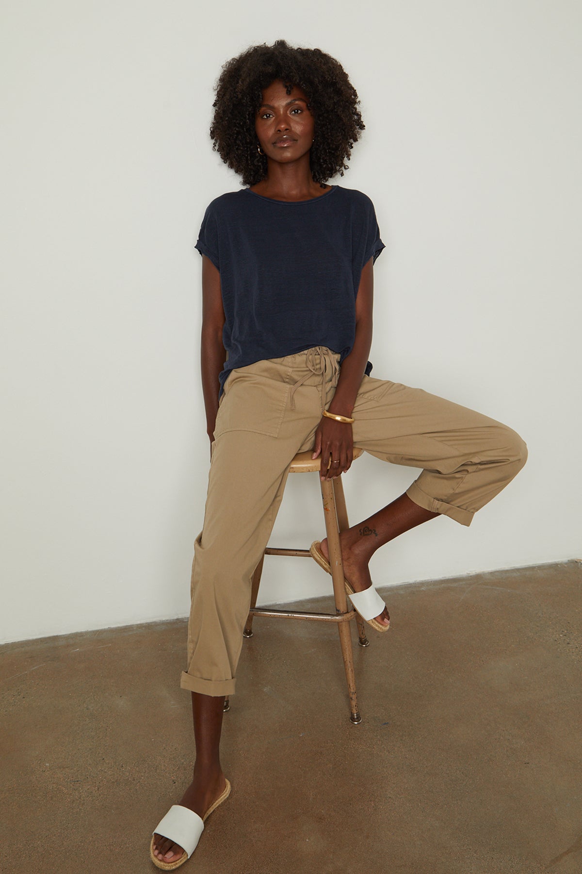   Woman sitting on stool in studio wearing Hudson tee in baltic blue with Misty pant in oak and white slides full length front 