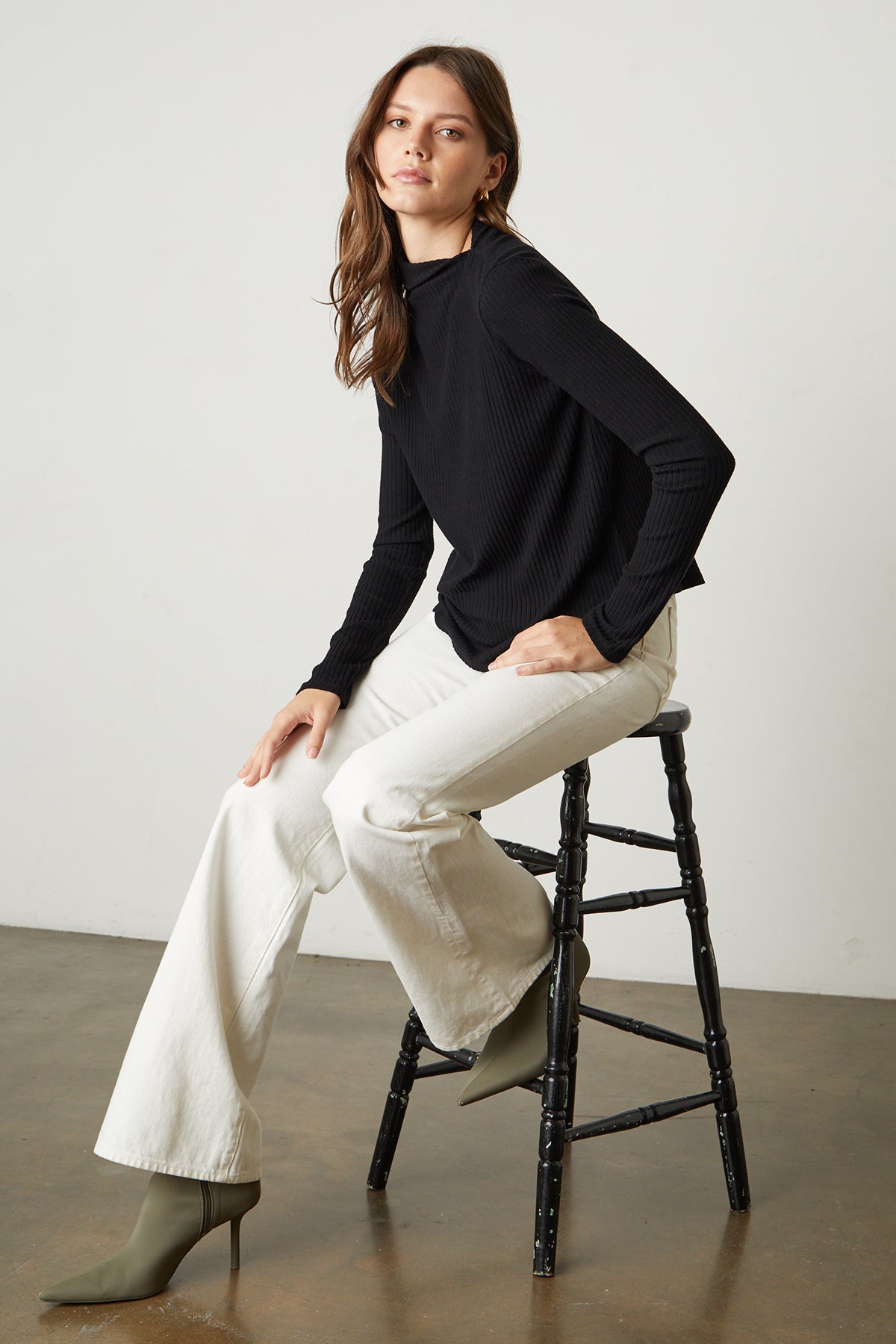   The model is sitting on a stool in a Velvet by Graham & Spencer DEANNA MOCK NECK TOP and white pants. 