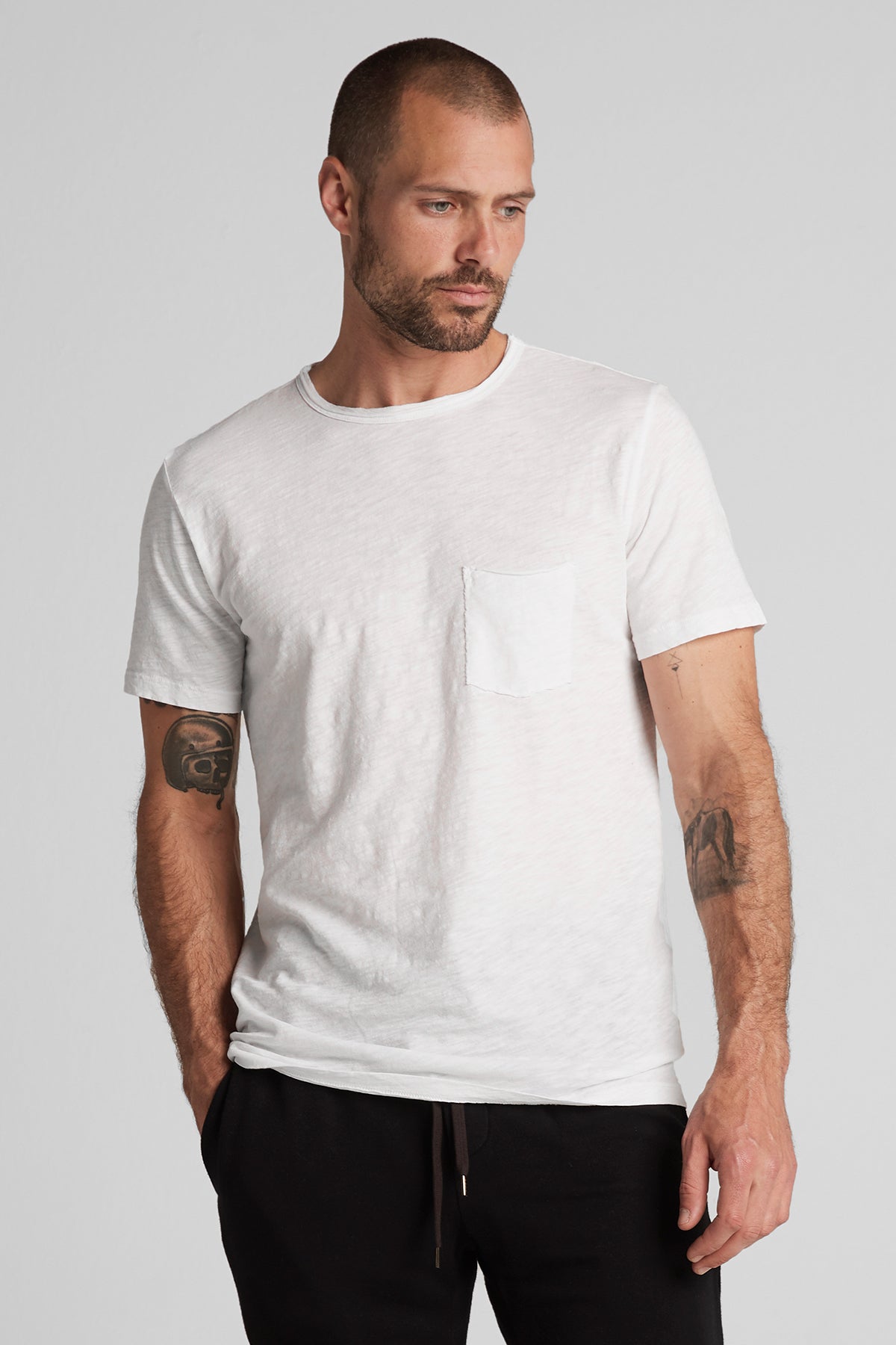 A man with tattoos on his arms, wearing a white textured cotton slub CHAD TEE with a chest pocket and black pants, stands against a neutral background. (Brand Name: Velvet by Graham & Spencer)-25485730709697