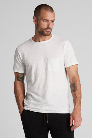 A man with tattoos on his arms, wearing a white textured cotton slub CHAD TEE with a chest pocket and black pants, stands against a neutral background. (Brand Name: Velvet by Graham & Spencer)