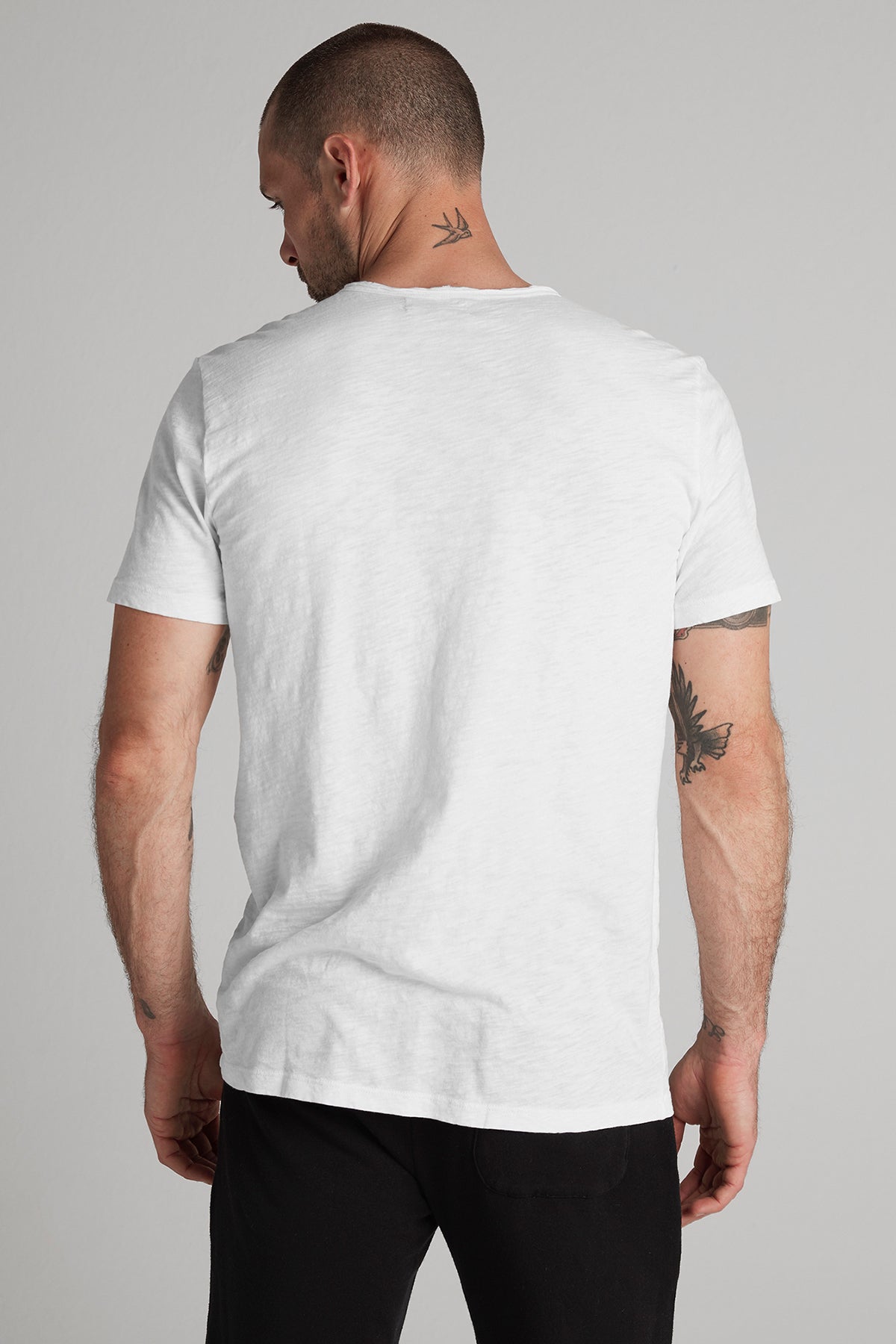   Man in a Velvet by Graham & Spencer CHAD TEE white t-shirt and black pants, viewed from behind, showing tattoos on both upper arms. 
