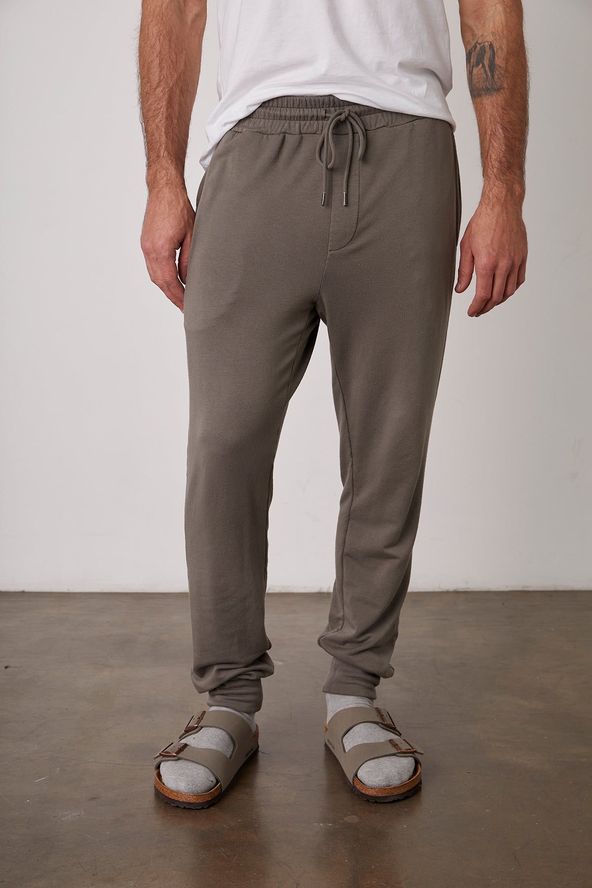   A person wearing a white t-shirt, the CROSBY LUXE FLEECE JOGGER by Velvet by Graham & Spencer, grey socks, and grey sandals stands indoors on a concrete floor. 