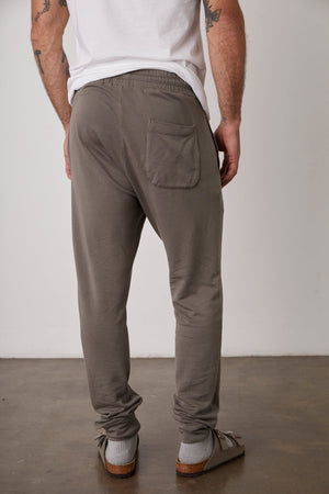 A person in a white shirt and gray sweatpants stands facing away, showcasing the back pocket of their CROSBY LUXE FLEECE JOGGER by Velvet by Graham & Spencer. They are wearing sandals with socks, standing on a concrete floor against a plain white wall.