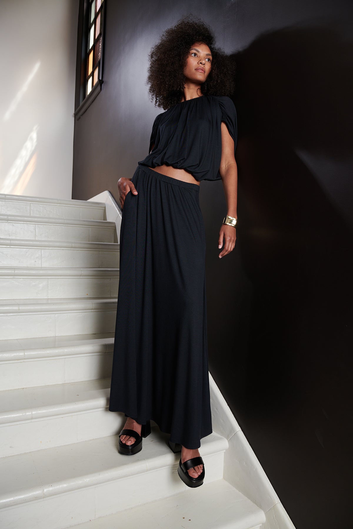 Model standing on steps with hand in pocket wearing Carmen Cocoon Drape Top in black with Malaya skirt in black.-25444320903361