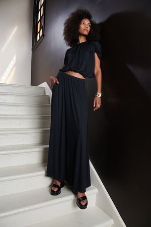 Model standing on steps with hand in pocket wearing Carmen Cocoon Drape Top in black with Malaya skirt in black.