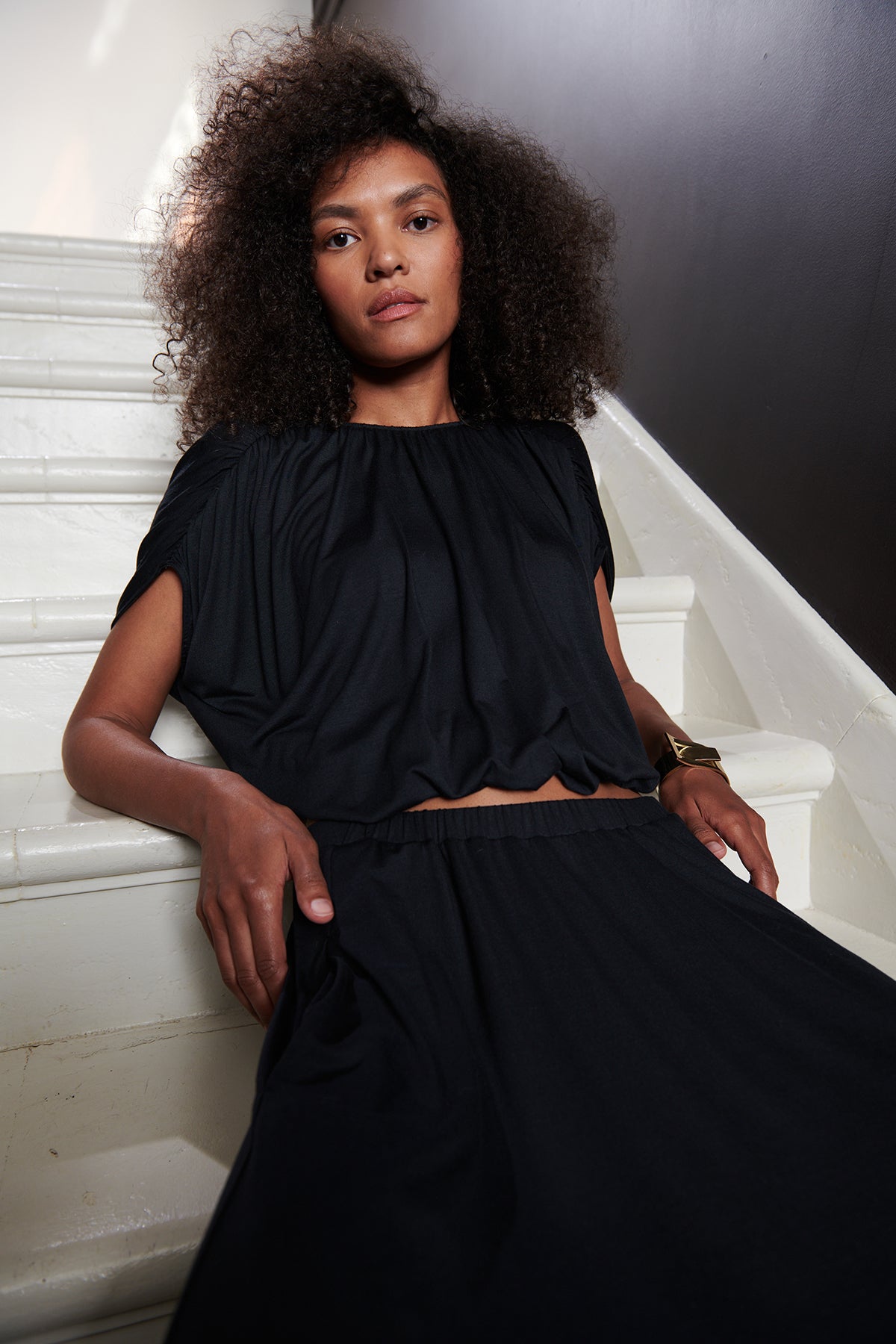 Model sitting on steps wearing Carmen Cocoon Drape Top in black with Malaya skirt in black.-25444318642369