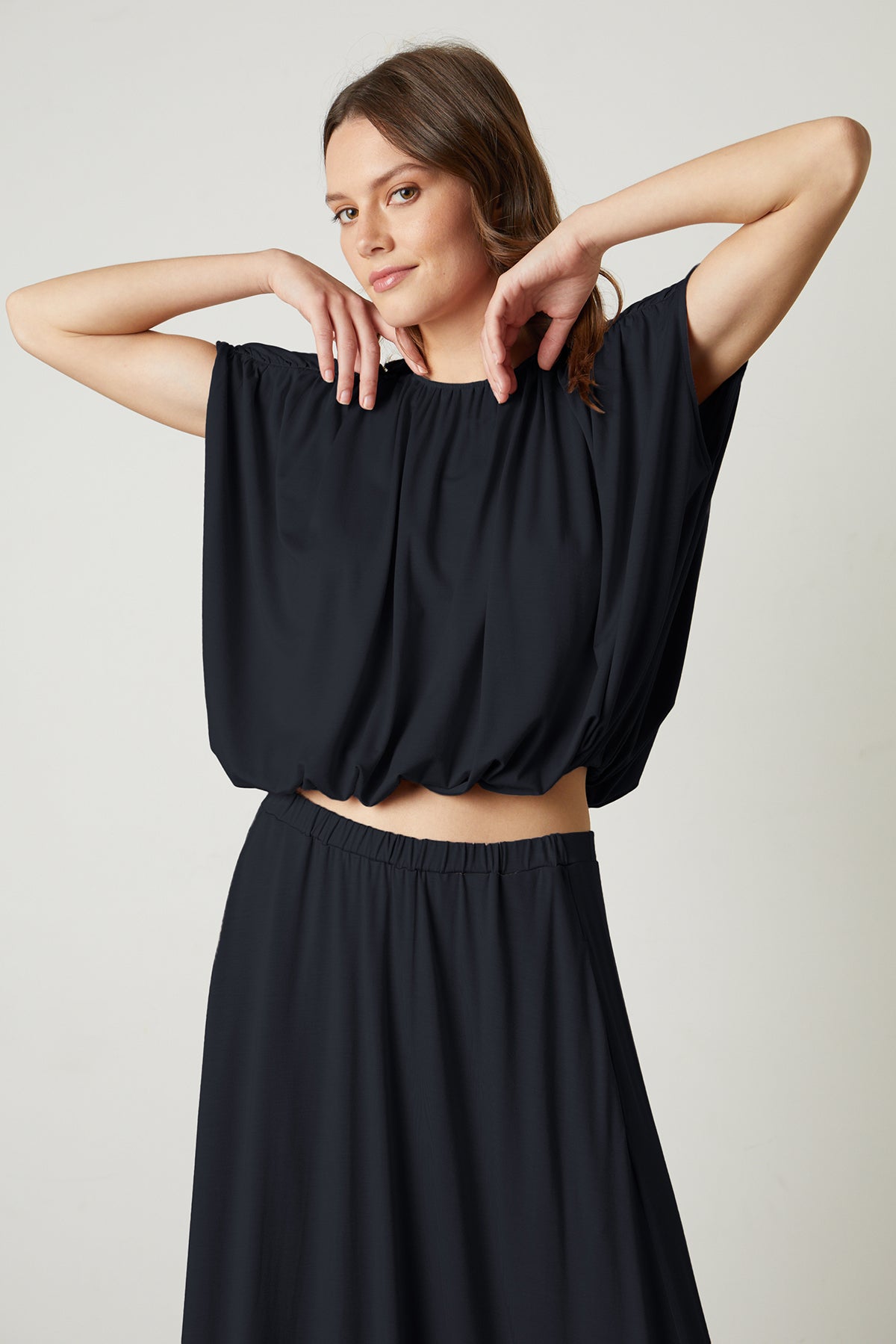   Model standing with arms up wearing Carmen Cocoon Drape Top in black with Malaya skirt in black. 