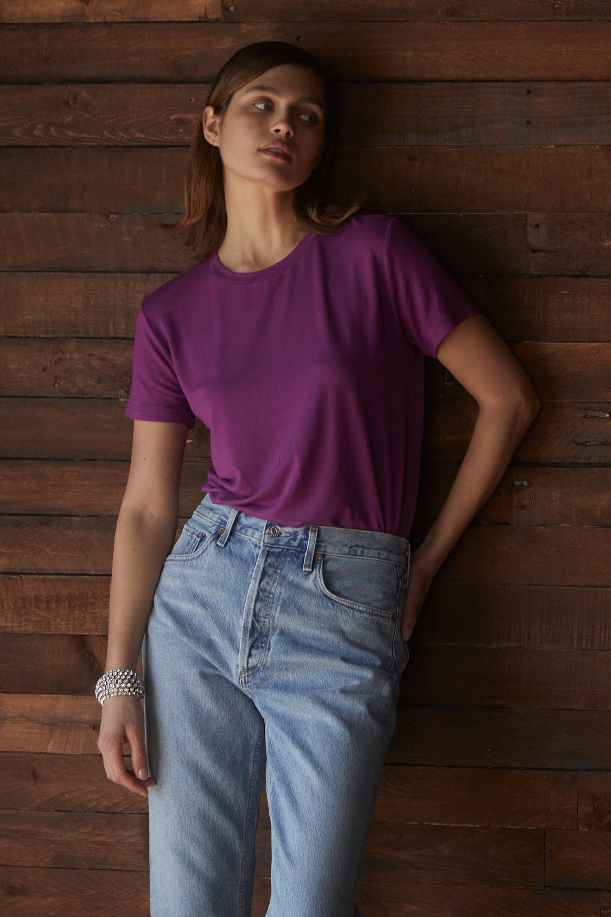   A woman in a tailored purple SOLANA TEE from Velvet by Jenny Graham leaning against a wooden wall with a soft modal jersey finish. 