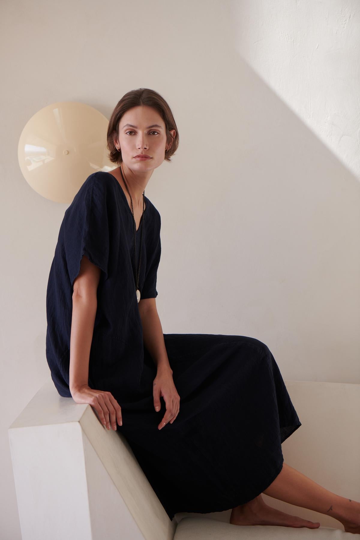 A woman is sitting on a white chair in a navy Velvet by Jenny Graham Montana dress.-26293194588353