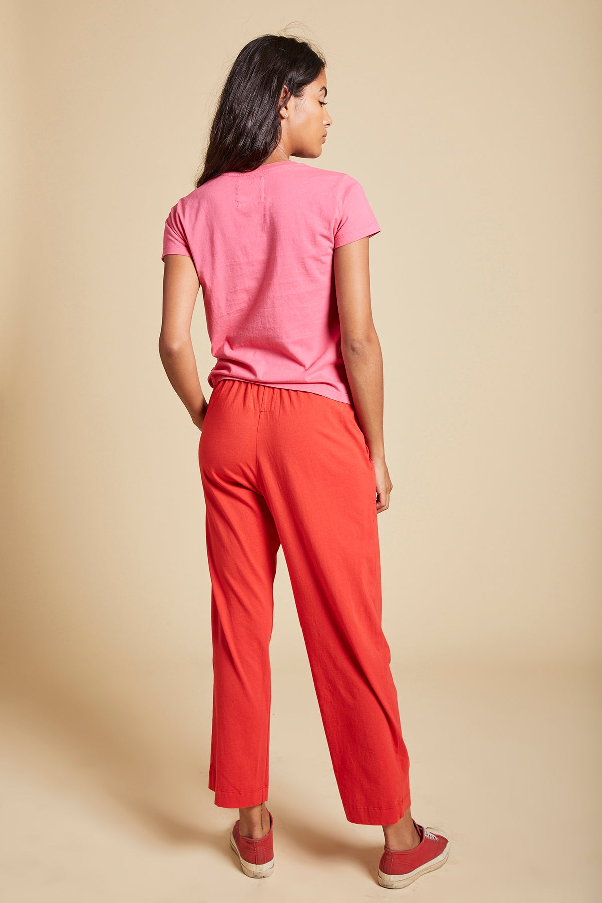   A person with long dark hair is standing with their back to the camera, wearing a pink t-shirt, PISMO PANT by Velvet by Jenny Graham, and red slip-on shoes, against a beige background. 