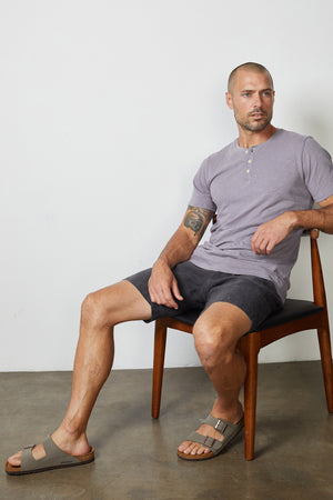 A man with a short beard sits on a wooden chair against a plain background. He wears a short-sleeve henley shirt, Velvet by Graham & Spencer's Maxwell Linen Shorts, and sandals—a perfect casual staple for relaxed days.