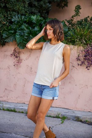 A person with short hair stands outdoors, wearing a tomboy-inspired sleeveless top called the TAURUS COTTON SLUB TANK by Velvet by Graham & Spencer, denim shorts, and brown shoes. They have one hand in their hair and are positioned in front of a pink wall with green foliage.