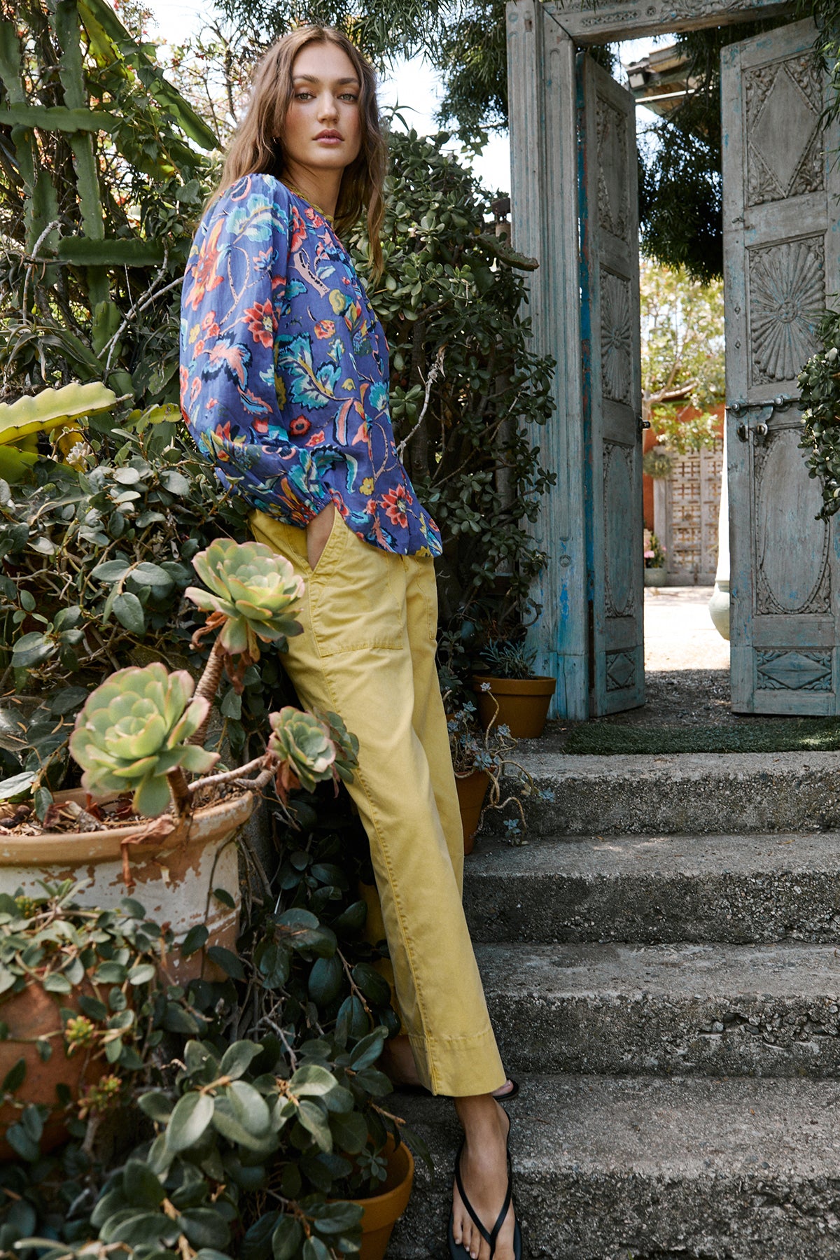   Model standing on garden steps outdoors wearing Camryn Top in colorful spring floral print with Mya pant in yellow aurora color side & front 