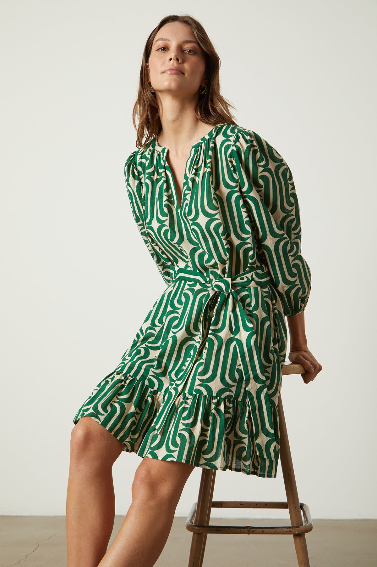   A woman is sitting on a stool wearing a FELICITY PRINTED BOHO DRESS by Velvet by Graham & Spencer with ruffled hemline. 